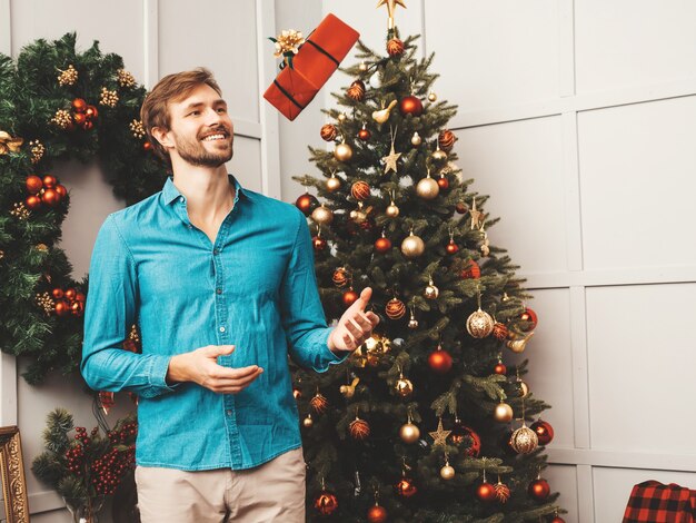 Retrato de sonriente hombre guapo con regalo. Hombre barbudo sexy posando cerca del árbol de Navidad con presente.