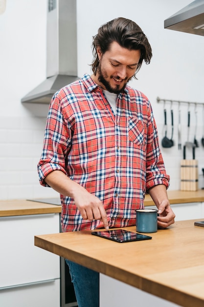 Retrato sonriente de un hombre hermoso que sostiene la taza de café que toca la pantalla de la tableta digital en la tabla