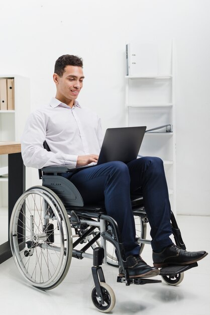 Retrato sonriente de un hombre joven discapacitado que se sienta en la silla de ruedas usando el ordenador portátil en el lugar de trabajo