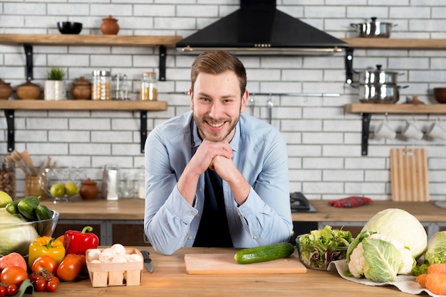 Foto gratuita retrato sonriente de un hombre joven que se coloca detrás de la tabla en la cocina