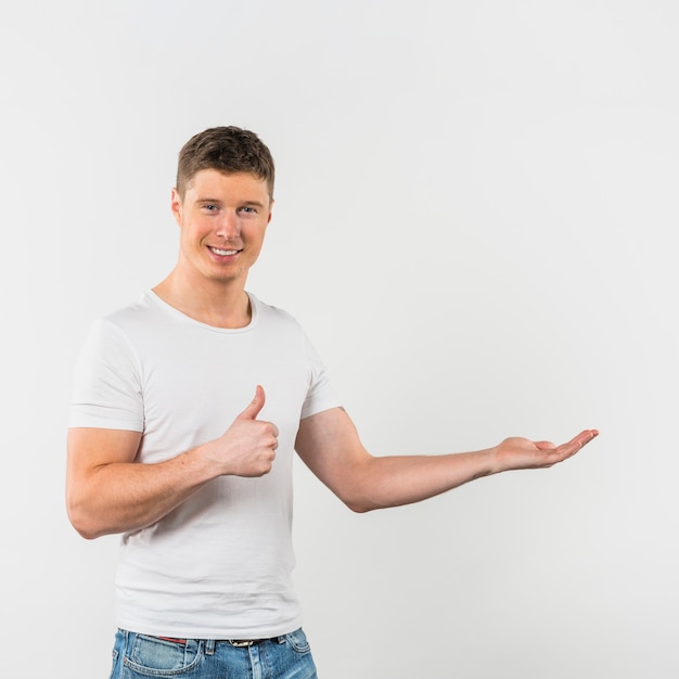 Retrato sonriente de un hombre joven que muestra el pulgar encima de la muestra que presenta contra el contexto blanco