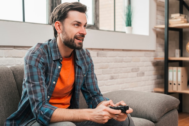 Retrato sonriente de un hombre joven que se sienta en el sofá que juega al videojuego