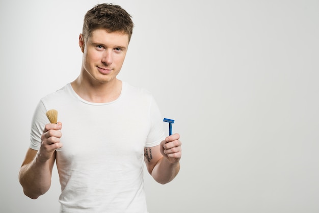 Retrato sonriente de un hombre joven que sostiene la maquinilla de afeitar y la brocha de afeitar en manos