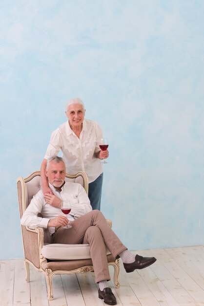 Retrato sonriente de un hombre mayor que se sienta en silla con su esposa que se coloca detrás de sostener el vidrio de vino