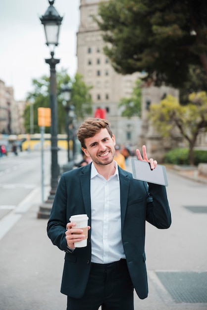 Retrato sonriente de un hombre de negocios que sostiene la taza de café para llevar y la tableta digital que muestran gesto de la victoria