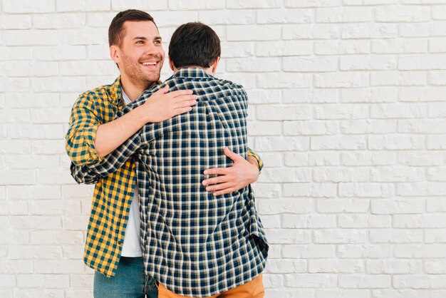 Retrato sonriente de un hombre que da un abrazo a su amigo contra la pared de ladrillo blanco