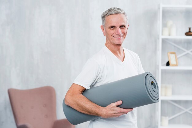 Retrato sonriente de un hombre que sostiene la estera rodada de la yoga en casa