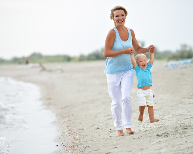 Retrato de sonriente joven madre con niño pequeño corriendo en la playa.