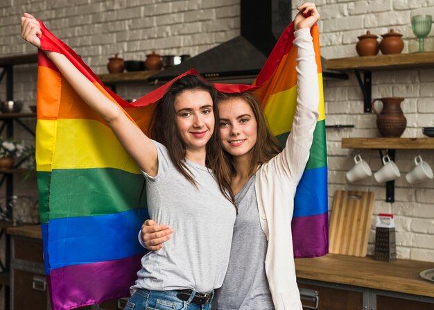 Retrato sonriente de una joven pareja de lesbianas sosteniendo la bandera del arco iris en la mano