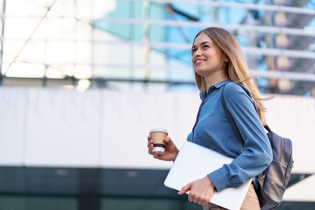 Retrato sonriente joven rubia sosteniendo el portátil y el café, vistiendo la camiseta azul suave sobre el edificio moderno