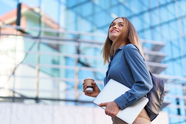 Retrato sonriente joven rubia sosteniendo el portátil y el café, vistiendo la camiseta azul suave sobre el edificio moderno