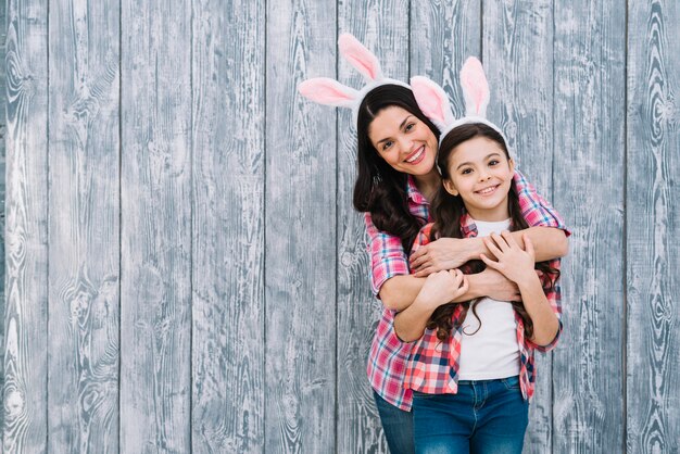 Retrato sonriente de madre abrazando a su hija por detrás frente a fondo de madera