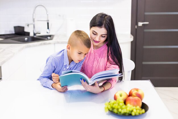 Retrato de sonriente madre ayudando a hijo con la tarea y pasar un buen rato en la cocina