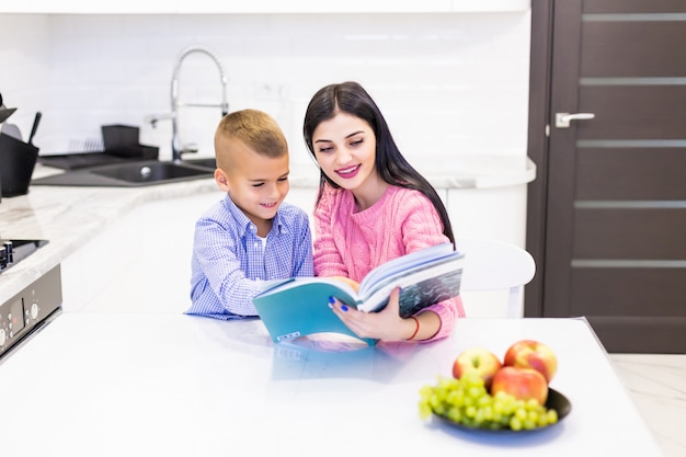 Retrato de sonriente madre ayudando a hijo con la tarea y pasar un buen rato en la cocina