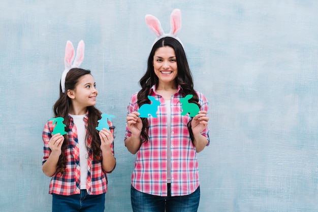 Retrato sonriente de la madre y la hija que sostienen el conejito de papel del recorte contra la pared azul