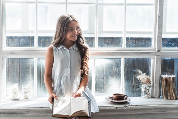 Retrato sonriente de una muchacha que se sienta en el travesaño de la ventana que sostiene el libro que mira lejos