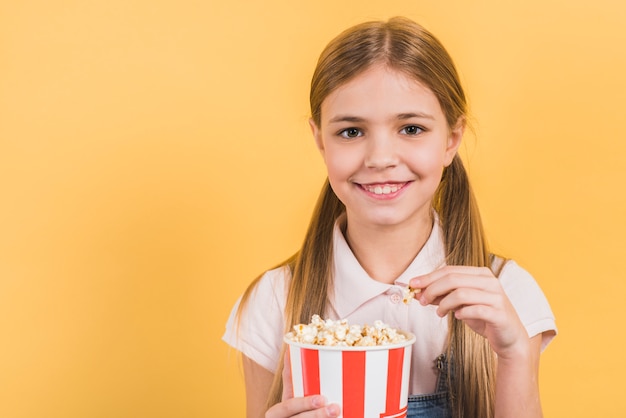 Retrato sonriente de una muchacha que sostiene el cubo de las palomitas contra el contexto amarillo