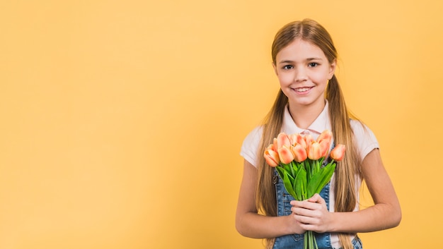 Foto gratuita retrato sonriente de una muchacha que sostiene tulipanes anaranjados disponibles contra el contexto amarillo