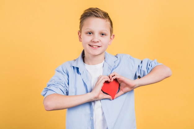 Foto gratuita retrato sonriente de un muchacho que muestra la forma roja del corazón que se opone al contexto amarillo