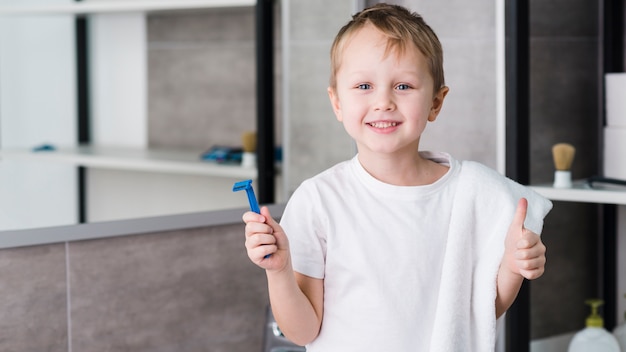 Retrato sonriente de un muchacho que sostiene la maquinilla de afeitar azul en la mano que muestra el pulgar encima de la muestra