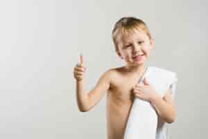 Foto gratuita retrato sonriente de un muchacho rubio descamisado con la toalla blanca en el hombro que muestra el pulgar encima de la muestra contra fondo gris