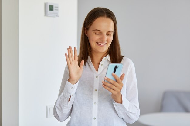 Retrato de sonriente mujer adulta joven de pelo oscuro con camisa blanca de pie en casa con el teléfono en las manos, con videollamada, agitando la mano a la cámara del dispositivo.