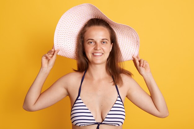 Retrato de sonriente mujer de cabello oscuro mujer con sombrero de paja y traje de baño blanco y negro a rayas con una sonrisa encantadora aislado sobre la pared amarilla.