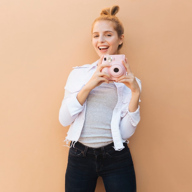 Retrato sonriente de una mujer hermosa joven que sostiene la cámara instantánea rosada contra el contexto beige