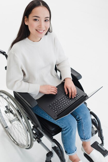 Retrato sonriente de una mujer joven que se sienta en la silla de ruedas con la computadora portátil en su regazo que mira la cámara