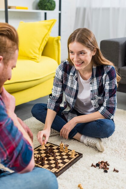 Retrato sonriente de la mujer joven que se sienta con su novio que juega a ajedrez en casa