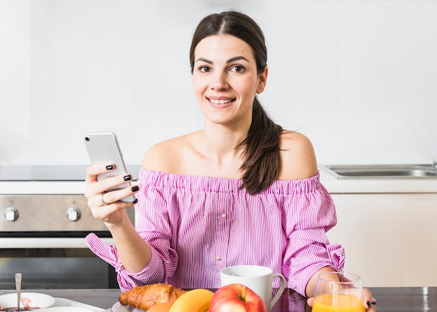 Foto gratuita retrato sonriente de una mujer joven que usa el teléfono móvil con el desayuno en la tabla