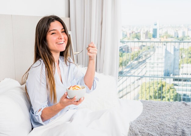 Retrato sonriente de una mujer joven sentada en la cama comiendo rodajas de manzana por la mañana