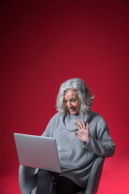 Retrato sonriente de una mujer mayor que agita su mano mientras que chatea video en la computadora portátil