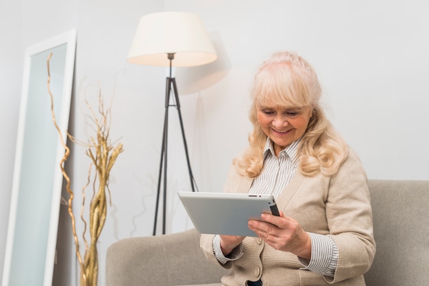 Retrato sonriente de una mujer mayor que se sienta en el sofá que mira la tableta digital