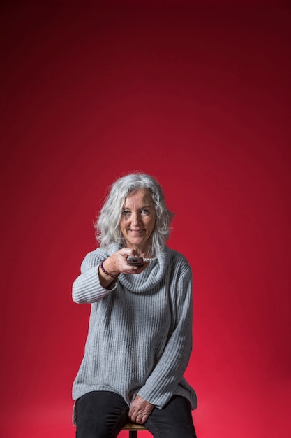 Retrato sonriente de una mujer mayor sentada en un taburete que cambia el canal con el control remoto contra el telón de fondo rojo