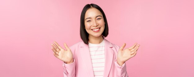 Retrato de una sonriente mujer de negocios de oficina asiática levantando las manos saludando y luciendo feliz posando en traje contra un fondo rosa