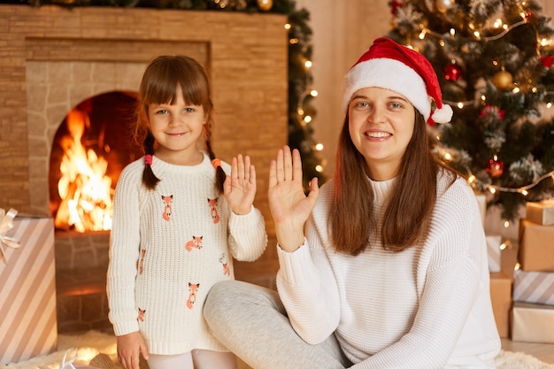 Retrato de sonriente mujer de pelo oscuro con suéter blanco y sombrero de santa claus posando con su pequeña hija, mirando a cámara y saludando con las manos, Feliz Navidad.