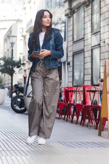 Retrato sonriente de una música que escucha de la mujer joven en el teléfono que se coloca en la calle