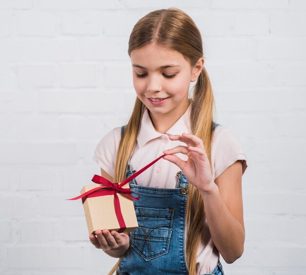 Retrato sonriente de una niña abriendo la caja de regalo de pie contra la pared de ladrillo blanco