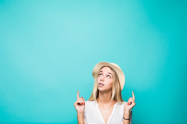 Retrato de una sonriente niña bonita de verano con sombrero de paja apuntando con dos dedos hacia arriba en copyspace aislado sobre la pared azul