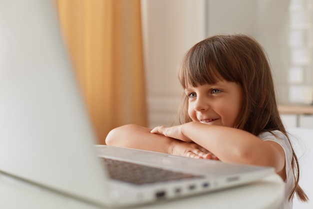 Retrato de sonriente niña pequeña de pelo oscuro sentada en la mesa frente a la computadora portátil y sonriente, niña en edad preescolar viendo dibujos animados o lección en línea.