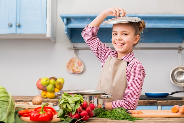 Retrato sonriente de una niña con tapa sobre su cabeza de pie en la cocina