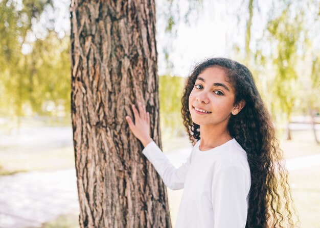 Retrato sonriente de una niña tocando el tronco de un árbol