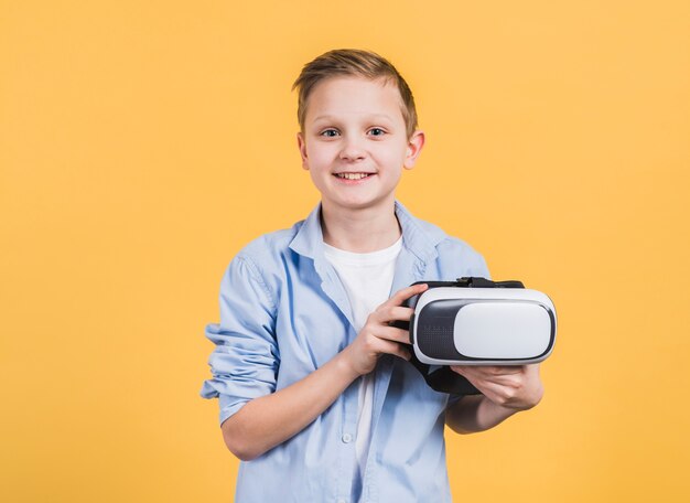 Retrato sonriente de un niño con gafas de realidad virtual en la mano contra el fondo amarillo