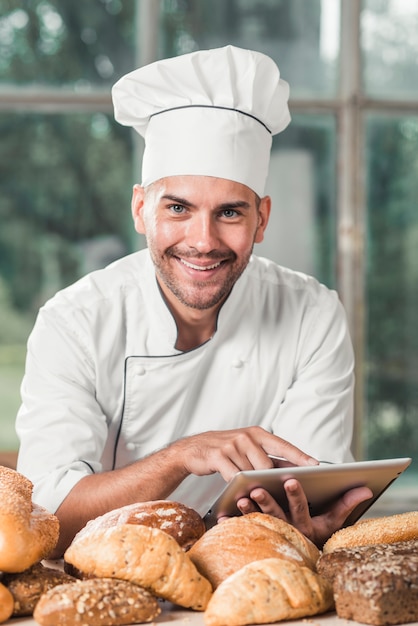 Retrato de sonriente panadero masculino usando tableta digital con muchos panes horneados