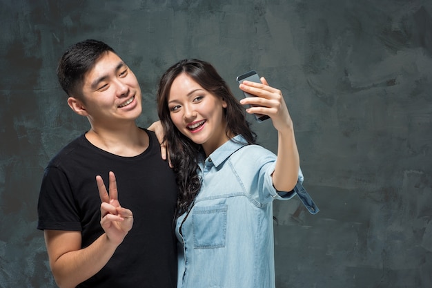 Retrato de sonriente pareja coreana haciendo foto selfie en un estudio gris