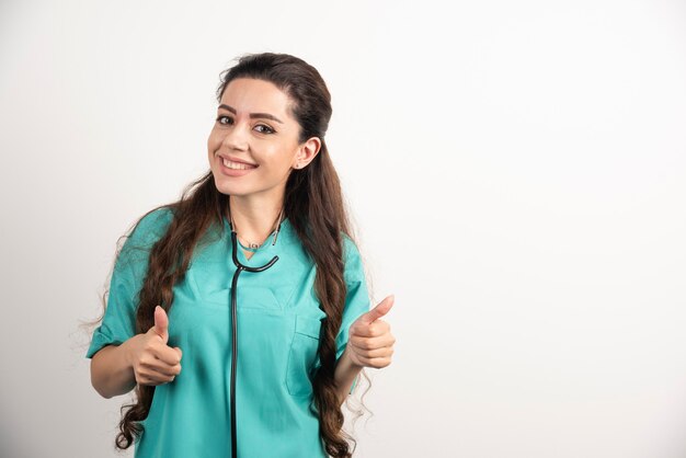 Retrato de sonriente trabajadora de la salud posando con el pulgar hacia arriba en la pared blanca.