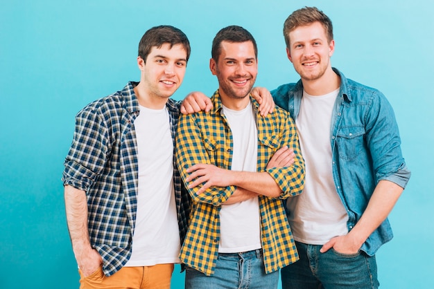 Retrato sonriente de tres amigos varones de pie contra el fondo azul