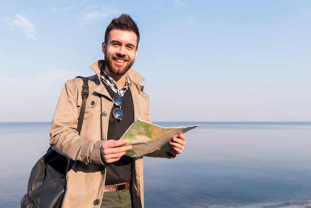 Retrato sonriente de un viajero masculino que se coloca delante del mar que sostiene el mapa disponible