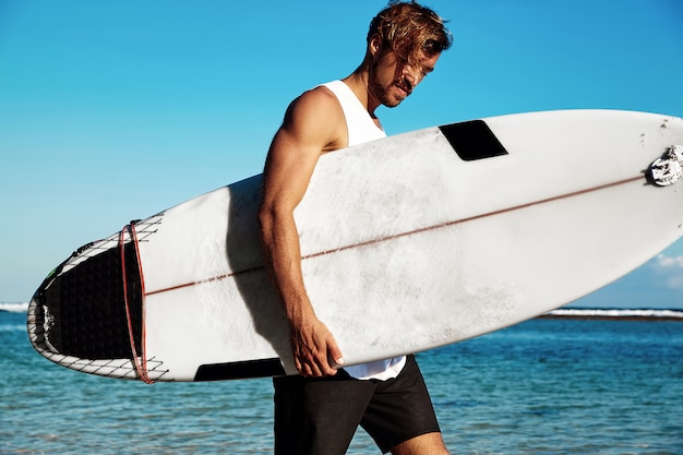 Retrato de surfista guapo modelo hipster tomar el sol moda hombre vistiendo ropa casual con tabla de surf en el océano azul y el cielo